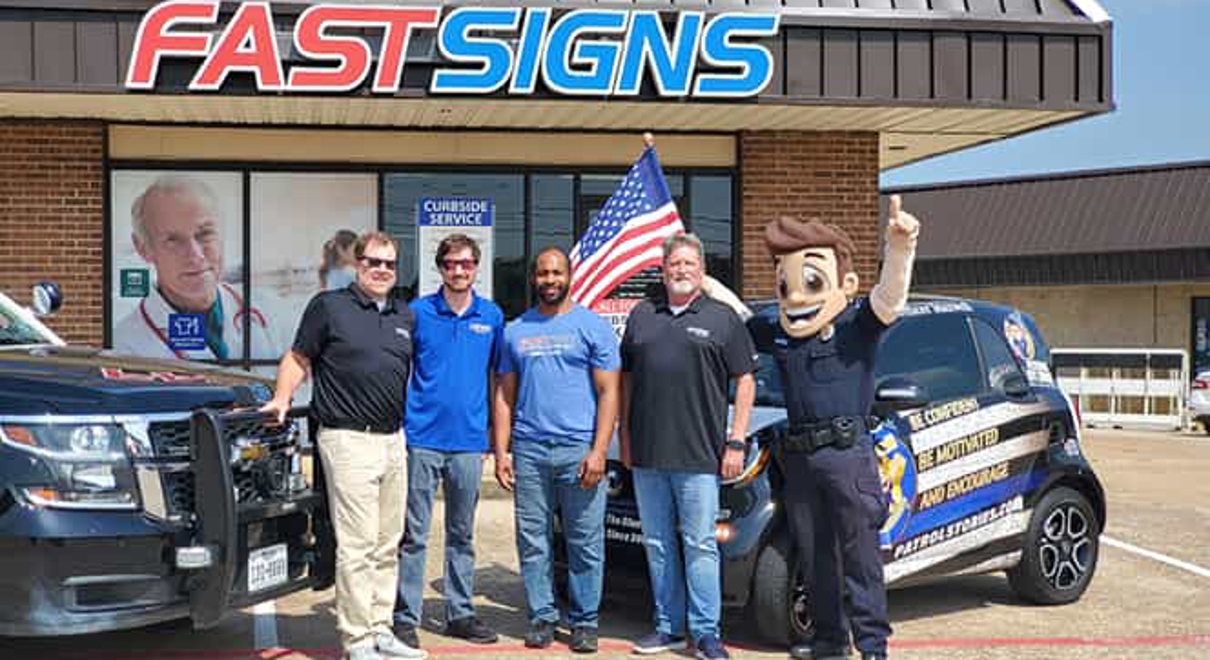A FASTSIGNS team poses out front with a wrapped vehicle and a mascot