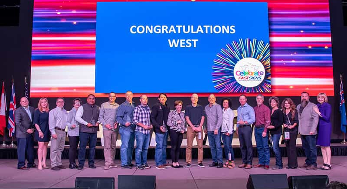 A FASTSIGNS team in front of a screen saying "Congratulations West"