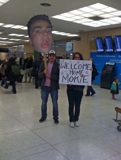 Two people hold a cutout of their friends face and a sign saying "Welcome Home Mokie"