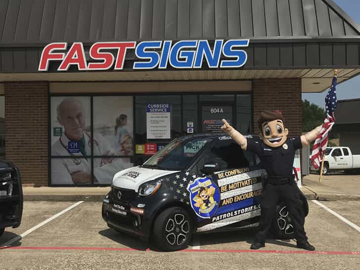 A cop mascot poses in a car with vehicle wrap outside of a fastsigns store