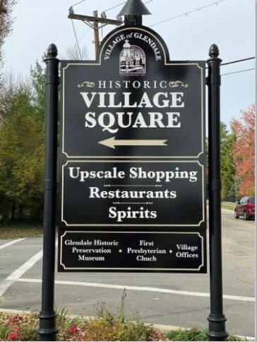 Village Square black signage in the street with trees, flower and red car