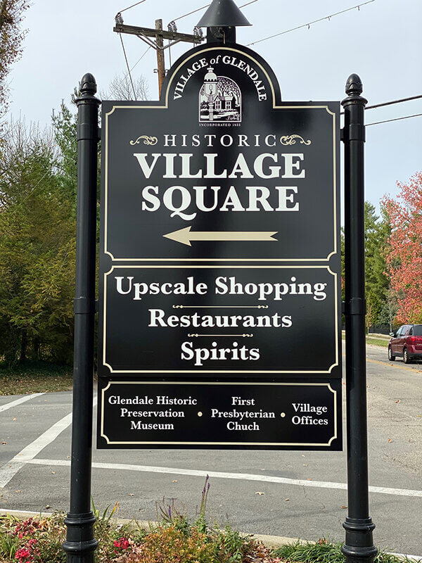Village Square black signage in the street with trees, flower and red car